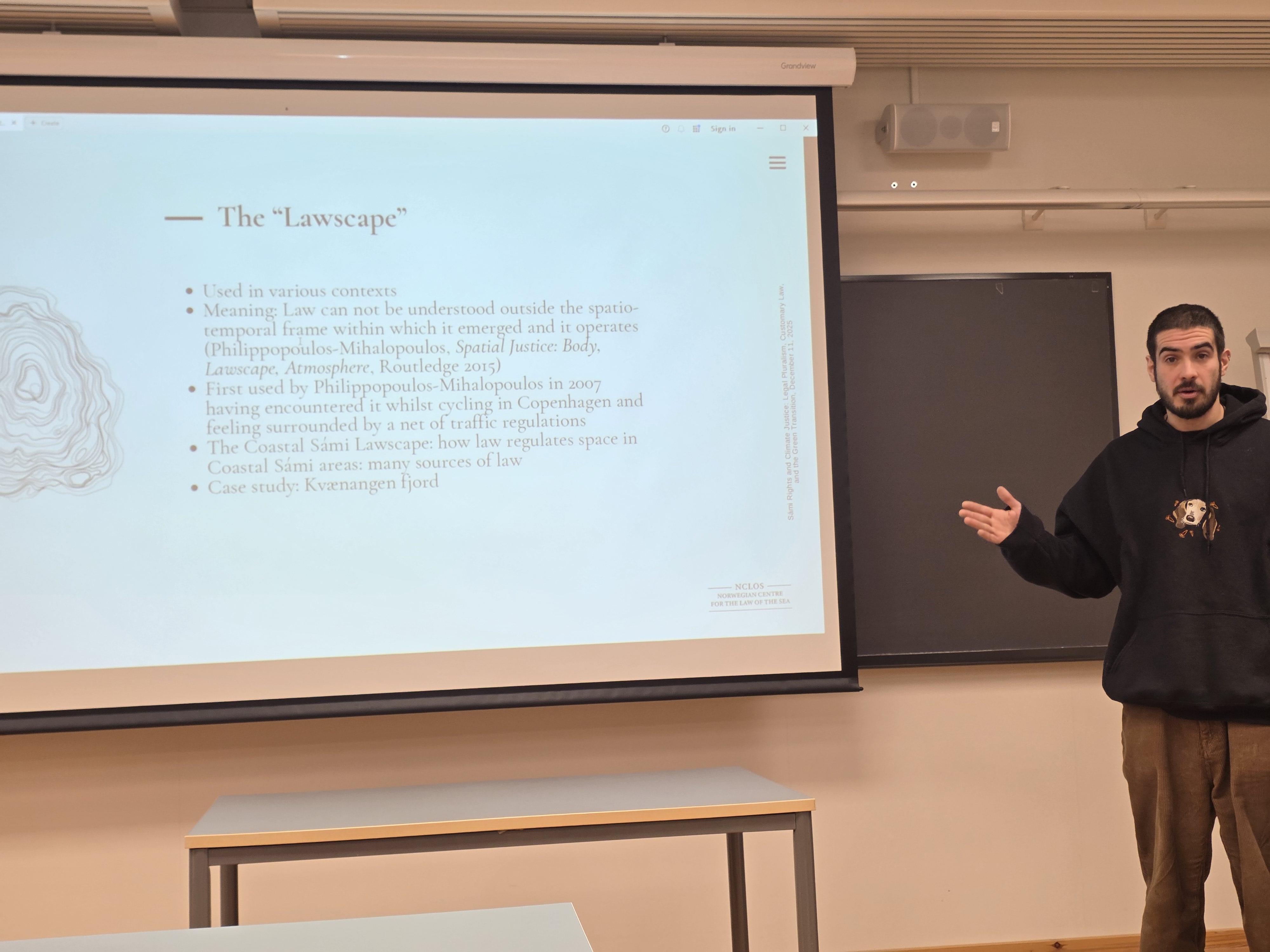 man is presenting to the audience, wearing a hoodie and brown pants, and holding his hand towards the powerpoint he is presenting with. 